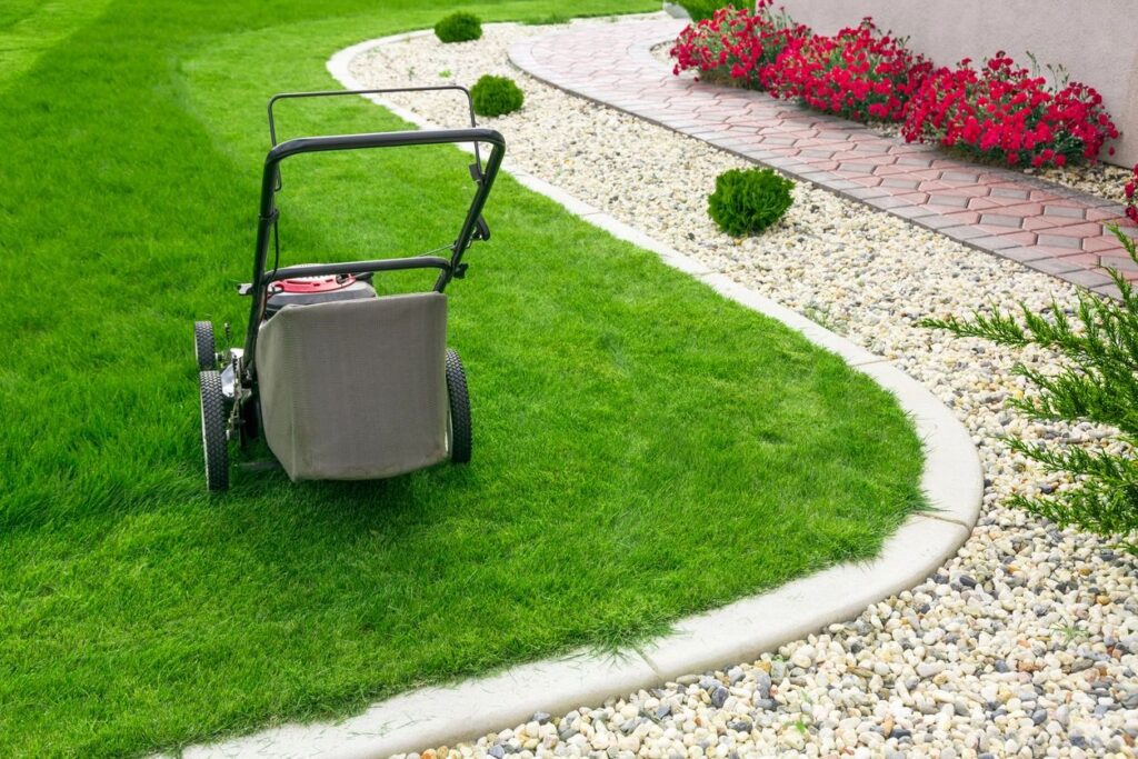 A lawn mower on a well-maintained grassy lawn beside a stone border and flowerbed with red flowers.