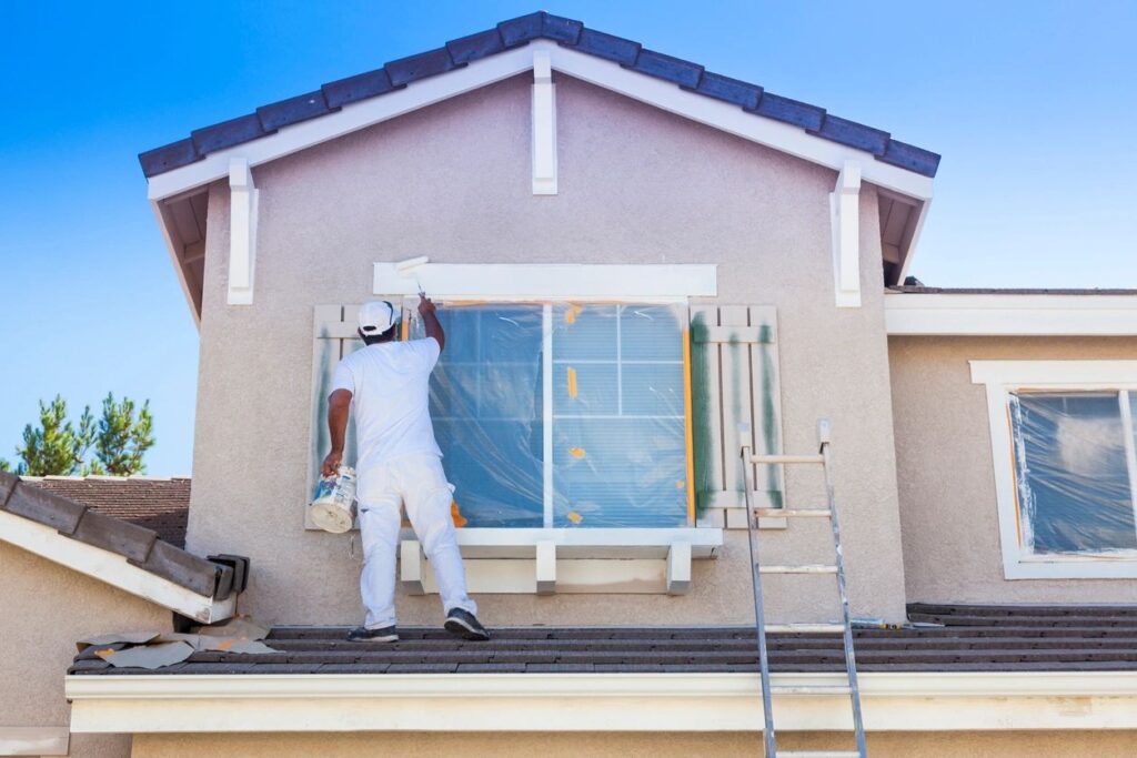 A worker in white painting the trim of a house window on a roof.
