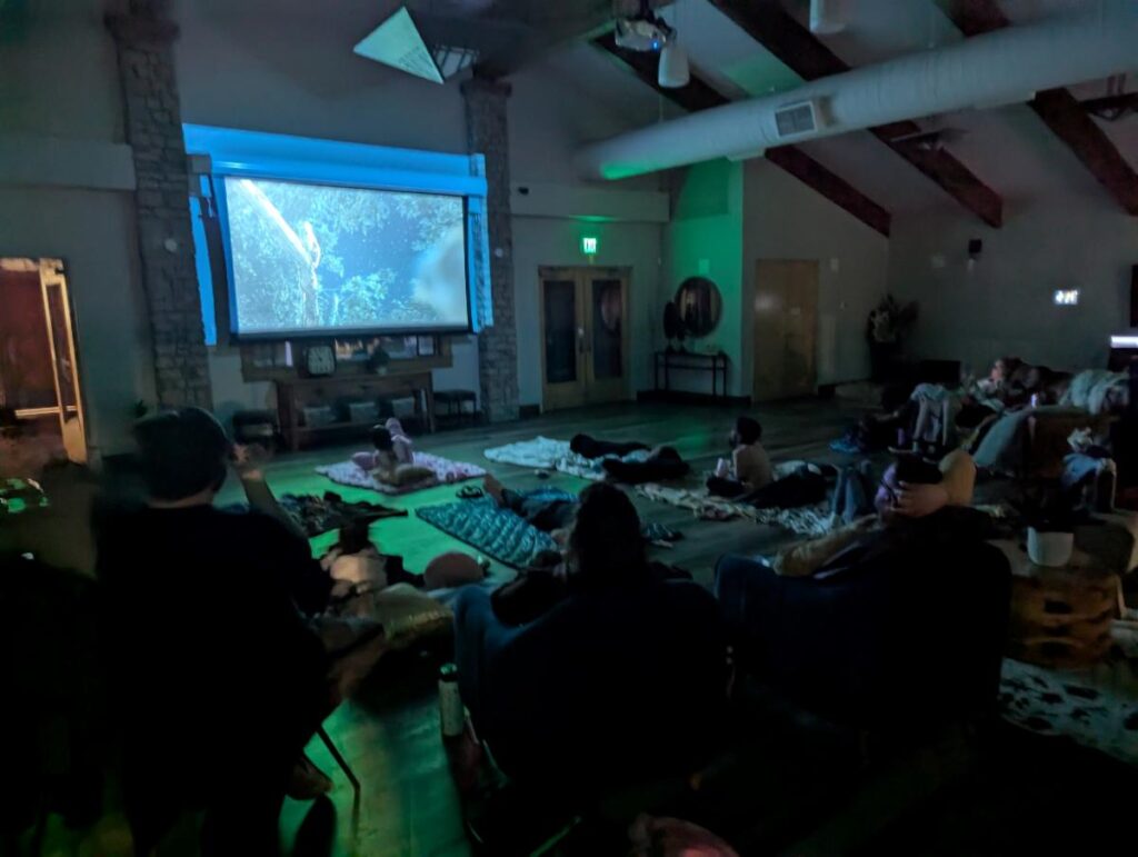 People watching a movie in a dimly lit room at the Wheatlands Clubhouse with a large screen and rustic decor.