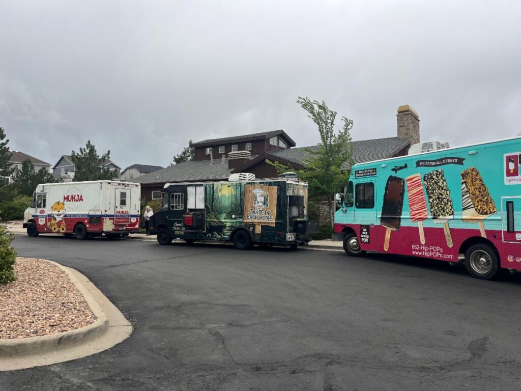 Three food trucks parked in front of the clubhouse with houses and cloudy sky in the background.