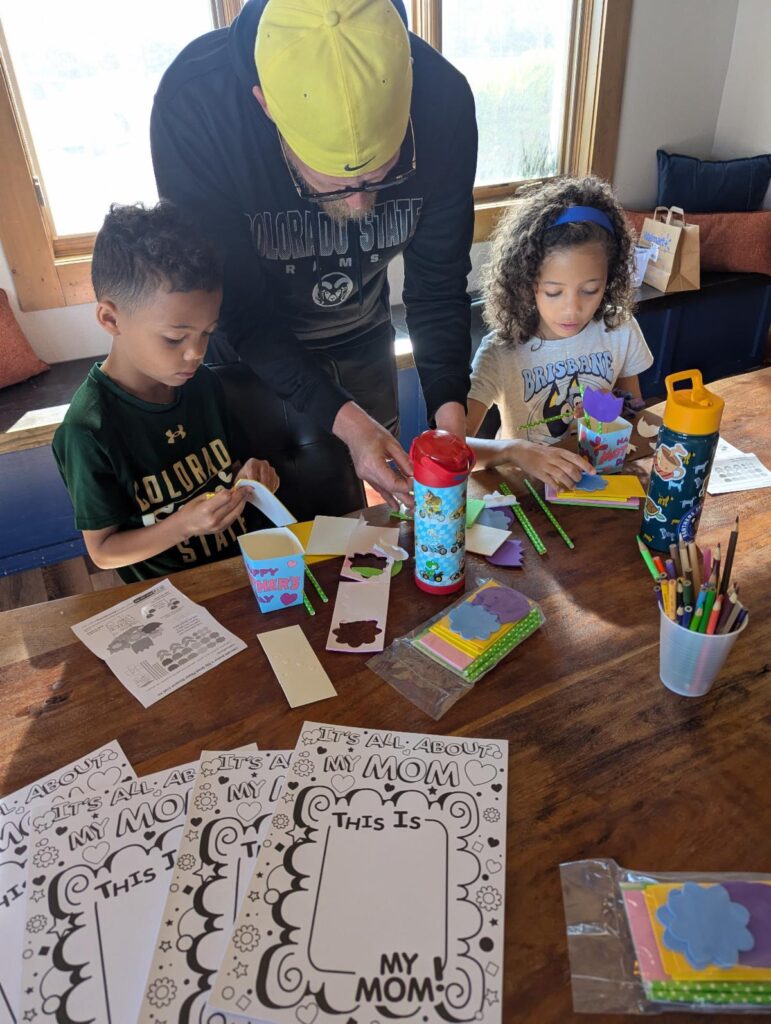 Two children and an adult male crafting at a table with Mother's Day-themed supplies.