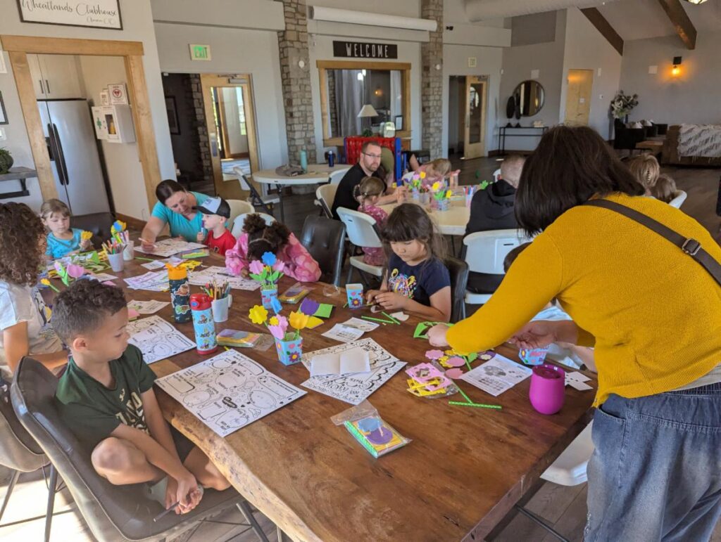 A group of children and adults engaged in a craft activity around a large wooden table with art supplies in a welcoming community room.