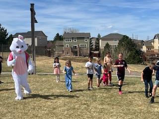 A person in a bunny costume leads children across a grassy field under a sunny sky.