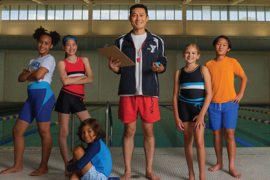 A male swim coach standing on the pool deck with a group of juvenile swimmers.
