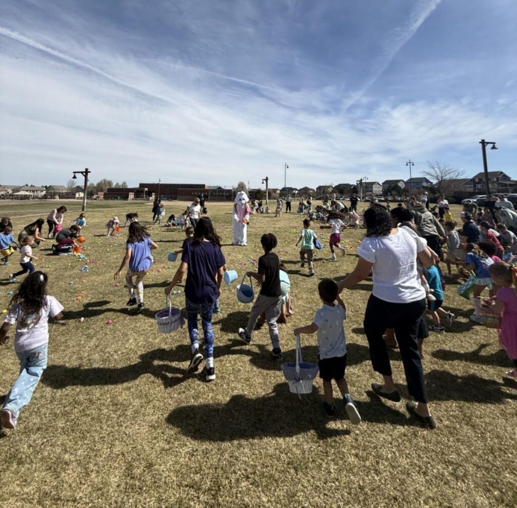 A large group of children and adults participating in an Easter egg hunt on a grassy field with a person in a bunny costume.