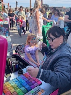 A face painter applying design to a young girl's face at an outdoor event with colorful paints.