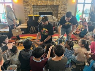 Children gathered around adults in a room with a fireplace, engaged in an activity.