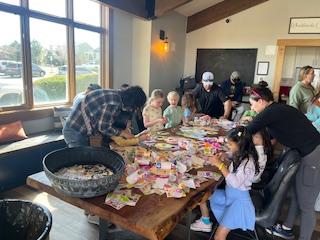 A group of people, including children, engaging in crafts at a large table in a well-lit room.